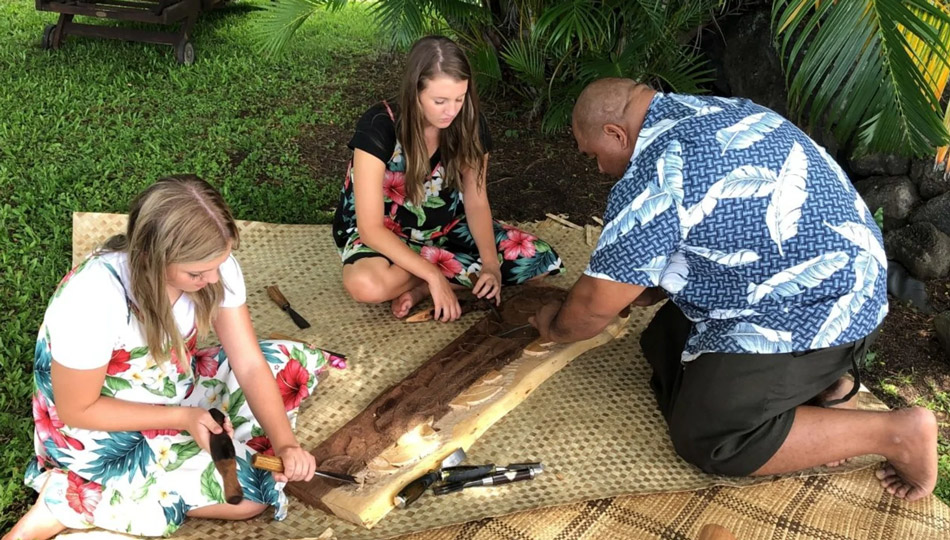 Polynesian woodworker carving a wood sculpture and teaching two young guests carving techniques